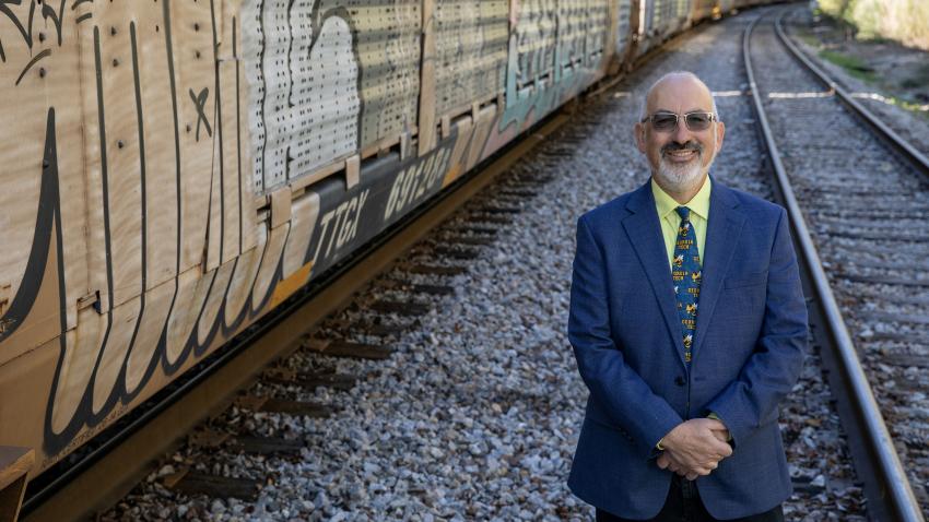 man wearing a blue blazer and a Georgia Tech themed tie stands with his arms crossed in front of him. He is smiling and wearing dark sunglasses. He is bald with a short white beard. He is standing next to train tracks on the right and a grafitti covered train car on the left.