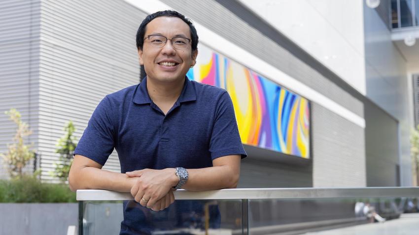 A smiling man standing outside. He is wearing a blue polo and is standing with his hands crossed in front of him on a railing. Behind him is a large screen with a colorful pattern.