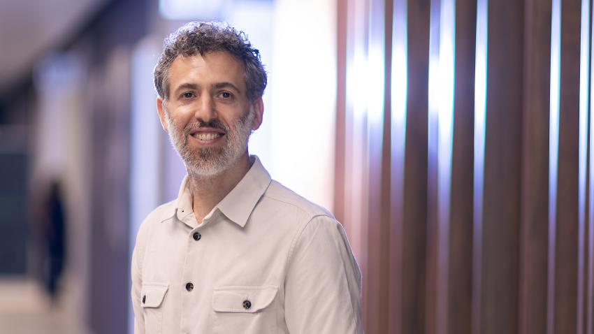 A man with salt and pepper hair and beard stands in a hallway wearing a white buton up shirt. There is a modern wooden panel behind him which reflects light and the purple color from the other walls