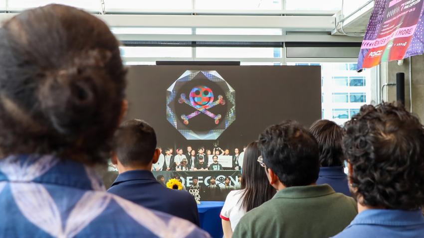 a croawd of students face a screen in the Coda Atrium. The screen shows a picture of the event stage from the hacking conference DefCon 33. On the stage is a crowd of people, including Team Atlanta. 