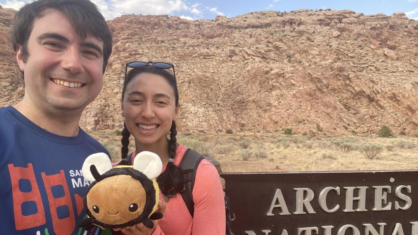 A man and a woman standing outside in the desert with a plush bee.
