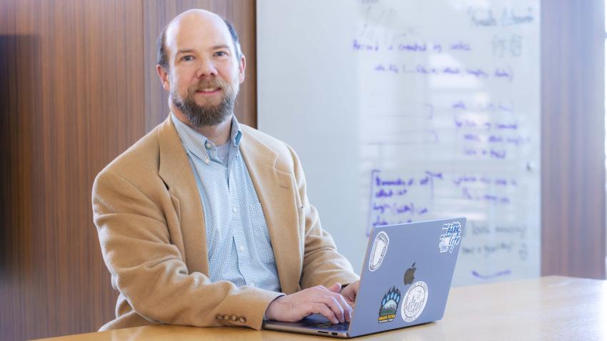 A man looks up from his computer and into the camera. Behind him is a white board with illegible writing on it. 
