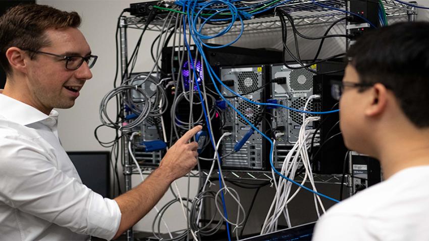 A man pointing to a rack of CPUs. There is a student to the right that is slightly out of focus. 