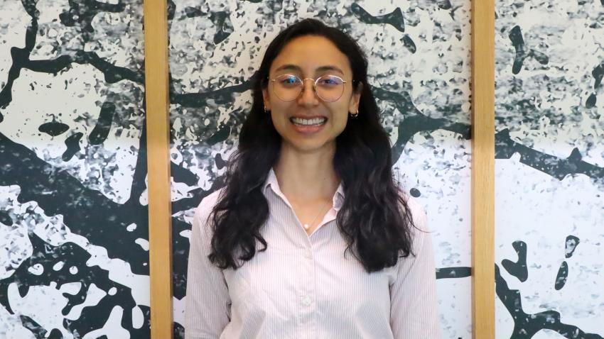 A smiling woman standing in front of a wall. The wall has a unique pattern and wooden frame.