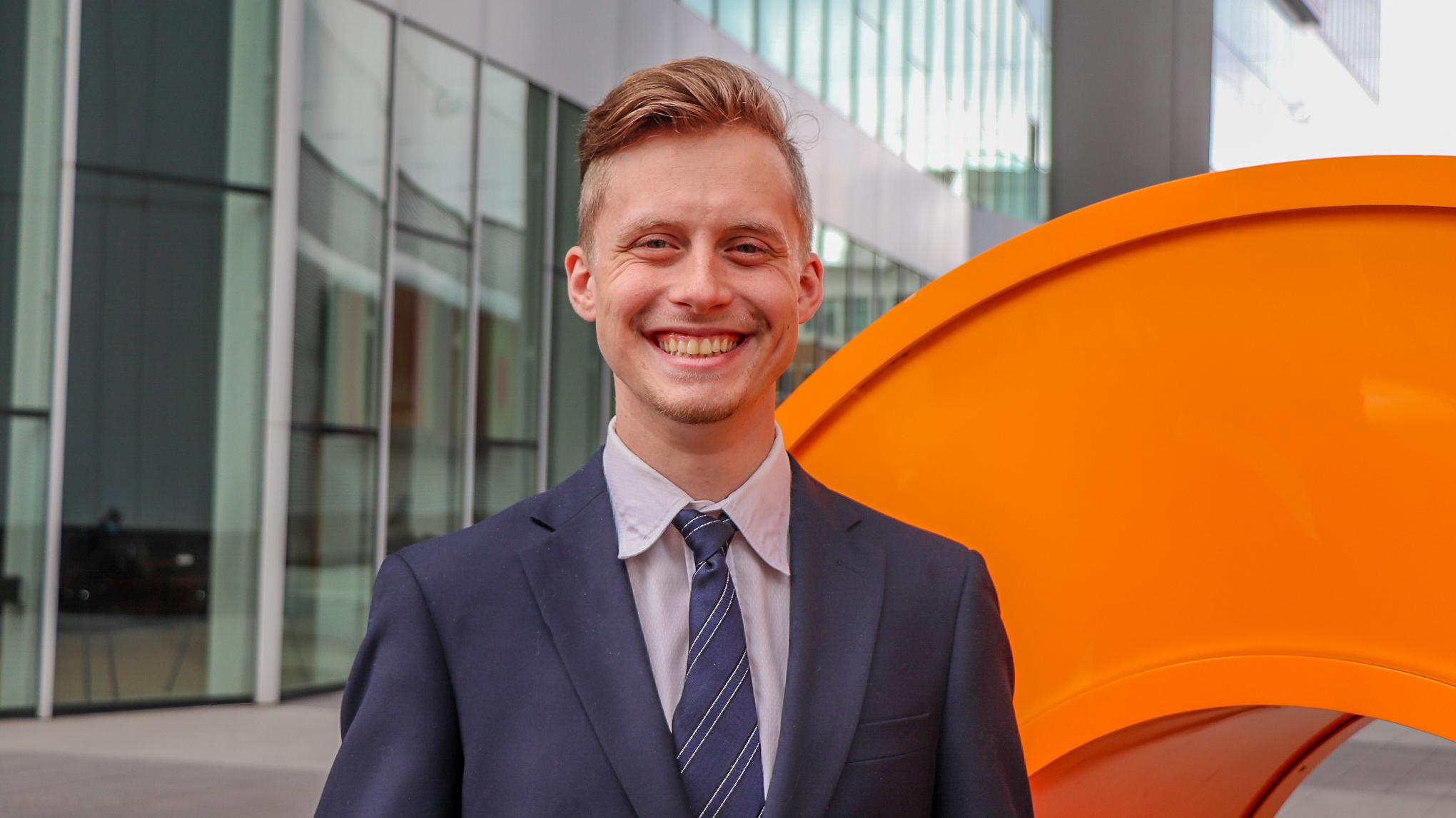 A man standing outside of the Coda Building in Tech Square. He is smiling and wearing a navy blue suit. 