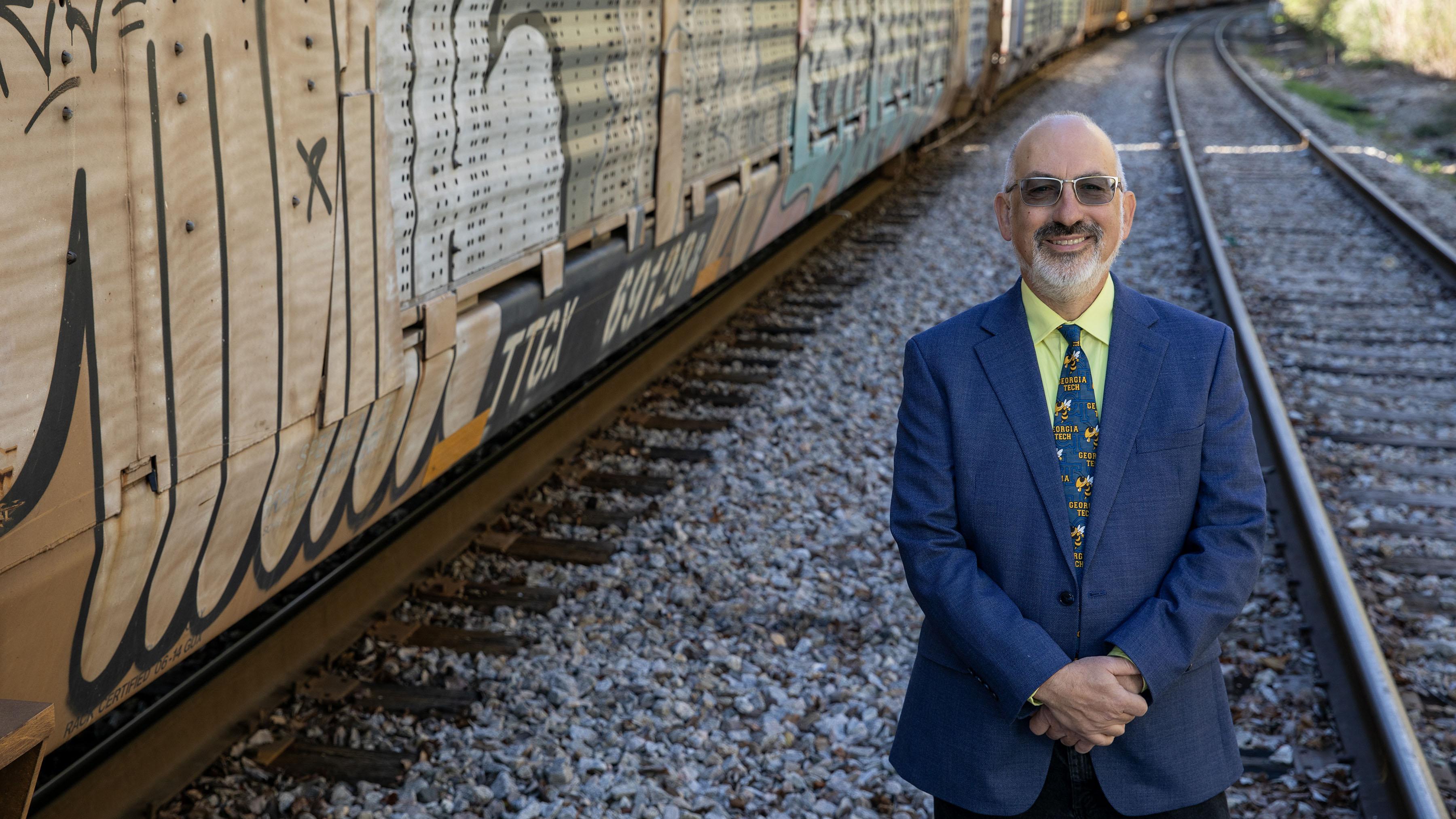 man wearing a blue blazer and a Georgia Tech themed tie stands with his arms crossed in front of him. He is smiling and wearing dark sunglasses. He is bald with a short white beard. He is standing next to train tracks on the right and a grafitti covered train car on the left. 