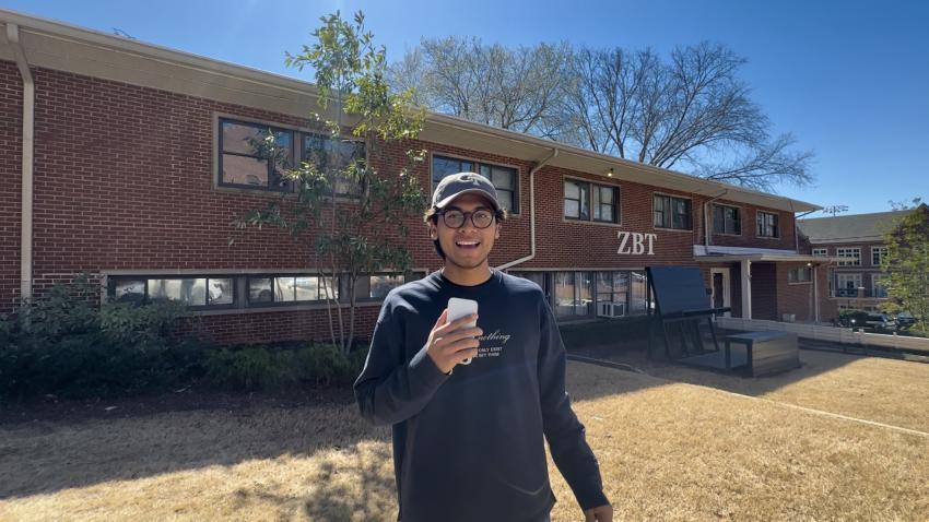 a young man wearing a ball cap stands in front of a long brick building. he has on glasses, a long sleeve navy shirt, and is smiling. He is holding up his smart phone as if to take a picture of the camera photographing him. 