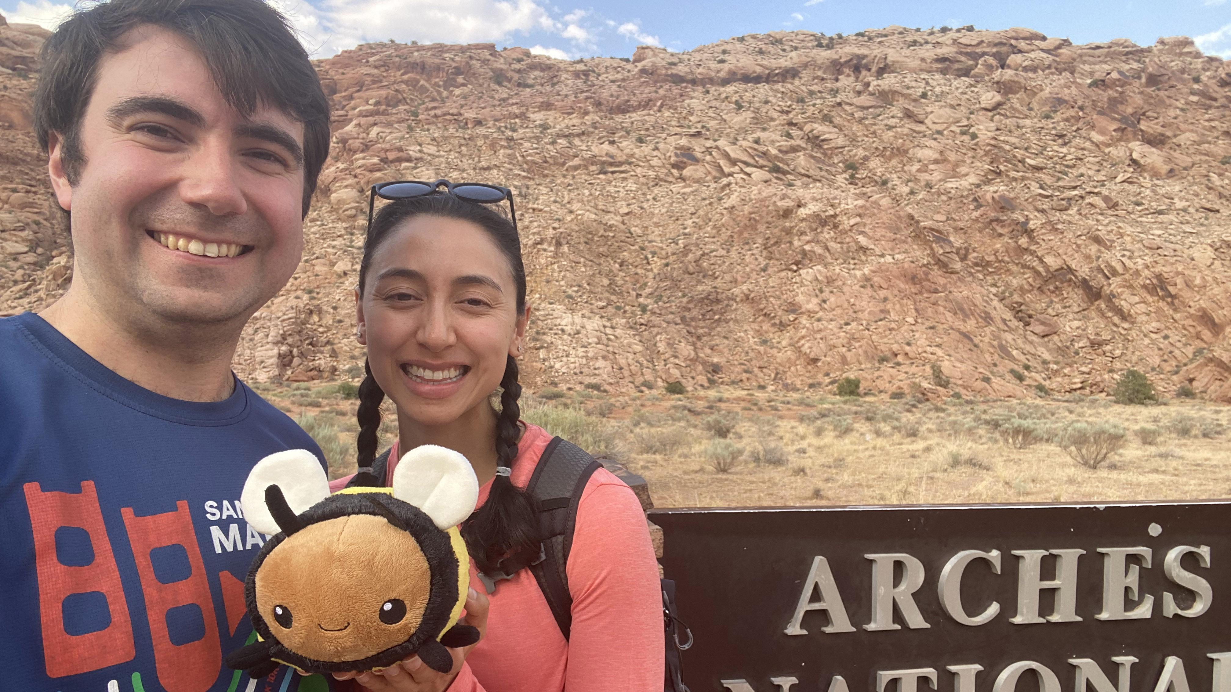 A man and a woman standing outside in the desert with a plush bee.