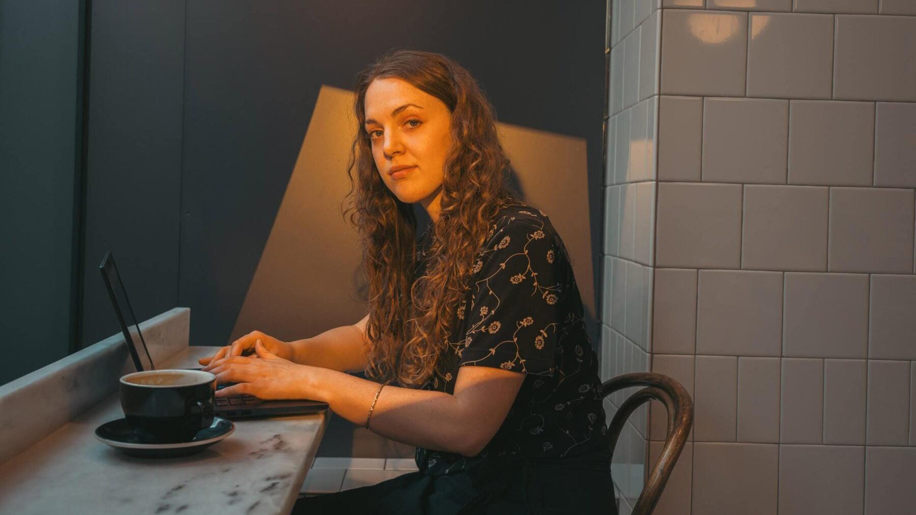 A young woman sitting at a counter with a laptop and a cup of coffee. She is looking into the camera with a serious expression on her face. She has long curly hair.