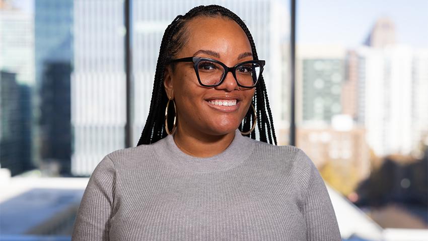 A smiling woman with braids wearing glasses and a grey sweater. 